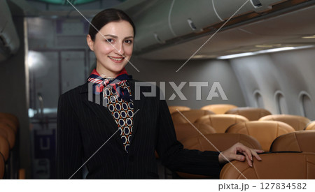 Smiling flight attendant in uniform welcomes and guides passengers aboard a modern airplane. 127834582
