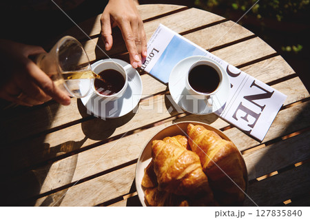 Glasses and cups of coffee with fresh croissants on a wooden outdoor table, bathed in soft sunlight, evoke a feeling of a relaxed morning or social cafe atmosphere. Glasses and cups of coffee with fresh croissants on a wooden outdoor table, bathed in soft sunlight, evoke a feeling of a relaxed morning or social cafe atmosphere. 127835840