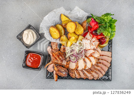 Assorted grilled meats served with roasted potatoes, pickled vegetables, fresh greens, and two dipping sauces on a black platter 127836249