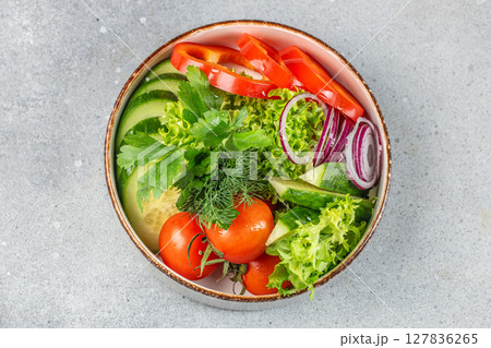 A fresh and colorful vegetable salad with cucumbers, tomatoes, red onions, bell peppers, lettuce, and herbs arranged in a rustic bowl on a gray stone surface 127836265