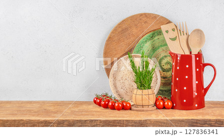 A cozy rustic kitchen setup with wooden utensils in a red polka dot jug, ceramic plates, cherry tomatoes, and fresh herbs arranged on a wooden counter against a light background 127836341