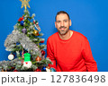 Bearded Hispanic man in his 40s wearing a red sweater posing satisfied and happy next to a Christmas tree, isolated on a blue studio background. Christmas concept. 127836498