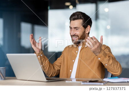 A businessman is happy and animated, with his hands raised in excitement, while participating in a video call using a laptop. 127837371