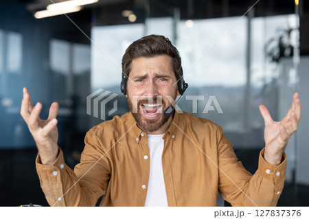 Businessman wearing headset looking frustrated handling a difficult customer support call. Professional man displaying feelings in tech office setting. Concept service communication. Businessman wearing headset looking frustrated handling a difficult customer support call. Professional man displaying feelings in tech office setting. Concept service communication. 127837376