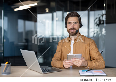 Smiling customer service representative wearing headset working remotely from modern office while holding tablet. Laptop on desk, showcasing professional technology use for communication. Smiling customer service representative wearing headset working remotely from modern office while holding tablet. Laptop on desk, showcasing professional technology use for communication. 127837461