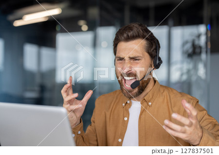 Businessman shows frustration with tech issues during work. Wearing headset, interacting with laptop. Challenges in professional environment, emotion of stress evident in expression. 127837501