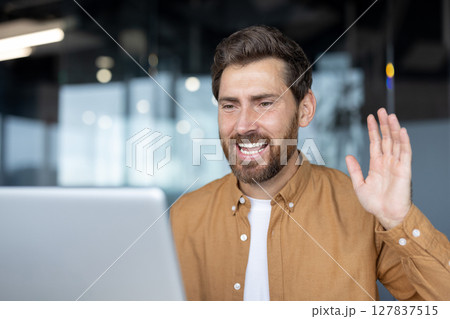 Man in brown shirt smiling during video call, greeting with raised hand in modern office space. Engaged in friendly online communication using laptop, conveying warmth and connection. 127837515