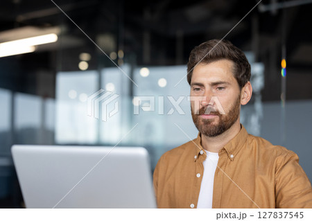 Man with beard using laptop in office. Reflects concentration, focus, productivity in modern workspace. Casual wear suggests informal but professional setting. Man with beard using laptop in office. Reflects concentration, focus, productivity in modern workspace. Casual wear suggests informal but professional setting. 127837545