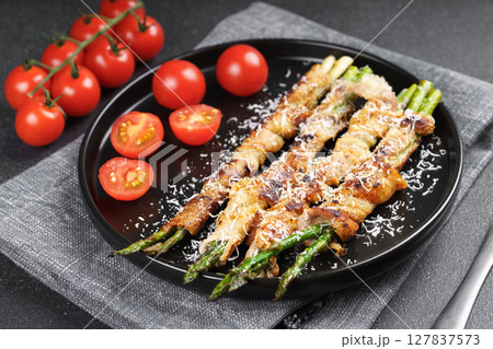 Grilled asparagus wrapped with bacon on black plate with cherry tomatoes on black background 127837573