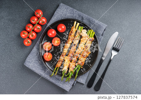 Top view of crispy bacon wrapped asparagus with cherry tomatoes on black plate and grey background 127837574