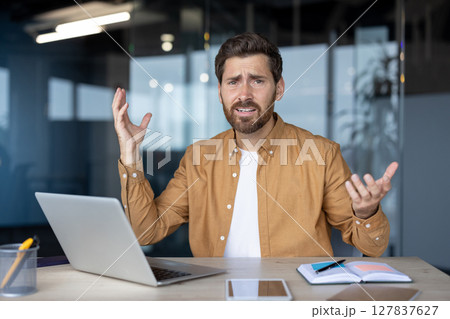 Frustrated businessman gestures at desk with laptop, notebook, phone, suggesting tech issue or confusion in office. Uncertainty and stress in workplace captured in moment. Frustrated businessman gestures at desk with laptop, notebook, phone, suggesting tech issue or confusion in office. Uncertainty and stress in workplace captured in moment. 127837627