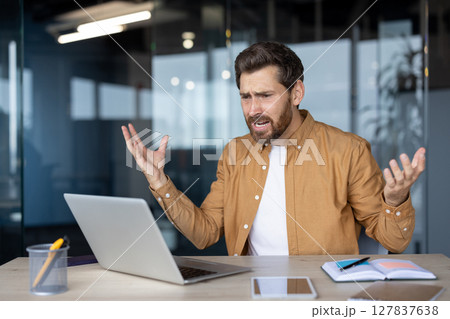 Mature businessman experiencing frustration at laptop in modern office. He gestures with raised hands, showing stress from remote work challenges or technical issues. Professional surroundings. 127837638