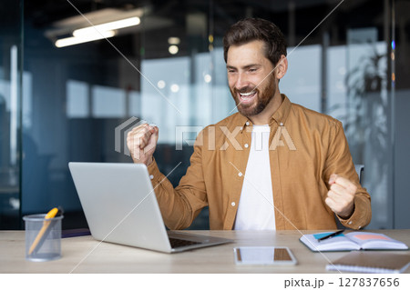 Mature business man jubilant over achievement indoors using laptop at desk. Expression of happiness and triumph. Office setting enhances feeling of professionalism and success. 127837656