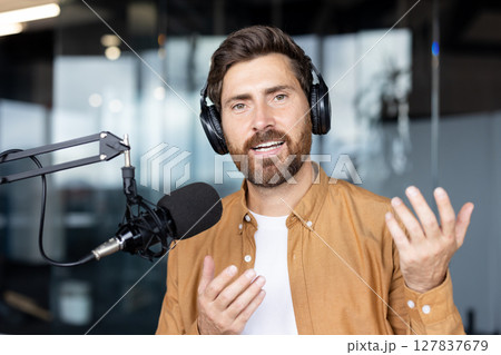 A man wearing headphones speaks into a microphone in a podcast studio setting, gesturing with his hands. 127837679