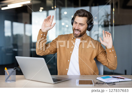 A man wearing headphones is happily gesturing in front of his laptop, possibly during a video call or listening to music. A man wearing headphones is happily gesturing in front of his laptop, possibly during a video call or listening to music. 127837683