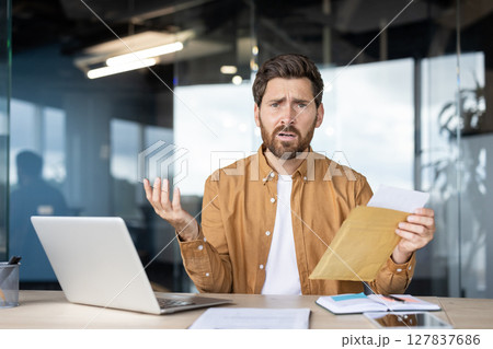 A concerned businessman holds an envelope with documents, looking perplexed at his office desk. A concerned businessman holds an envelope with documents, looking perplexed at his office desk. 127837686