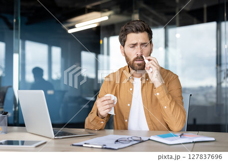 A man at his office desk is experiencing a cold, using a tissue to blow his nose while working. A man at his office desk is experiencing a cold, using a tissue to blow his nose while working. 127837696