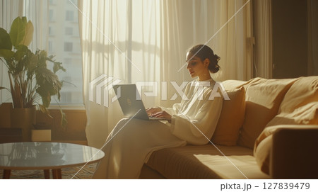 Young woman sitting on a comfortable couch in her living room, using a laptop and bathed in warm sunlight filtering through the window, creating a peaceful and productive work from home environment 127839479