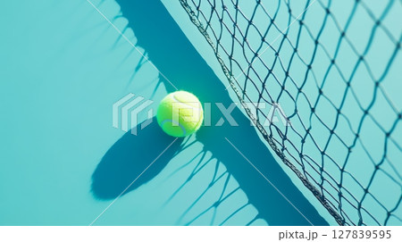 Vibrant yellow tennis ball lies near the net on a turquoise court, casting a long shadow under the bright sunlight, creating a dynamic composition that embodies the spirit of the game 127839595