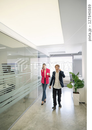 Two colleagues are walking through the office and talking on the phones.  127840889