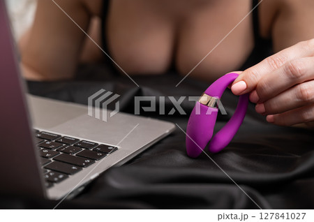 A woman is lying in bed holding a curved vibrator and looking at a laptop. A woman is lying in bed holding a curved vibrator and looking at a laptop. 127841027