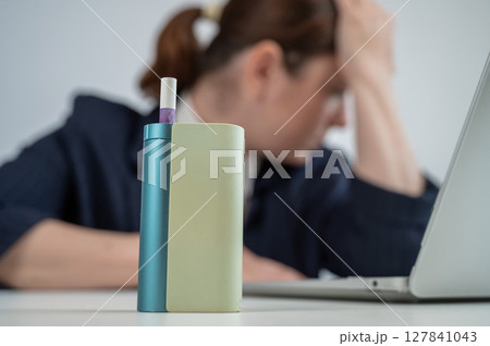 Tired businesswoman smoking an electronic cigarette while working on a laptop. Tobacco heating system. Tired businesswoman smoking an electronic cigarette while working on a laptop. Tobacco heating system. 127841043