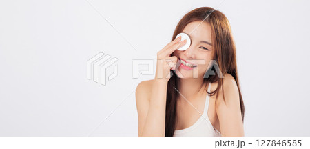 Pad in hand and cotton pad on cheek as an Asian woman removes makeup, revealing clean skin and natural beauty with a gentle smile. isolated studio white background 127846585
