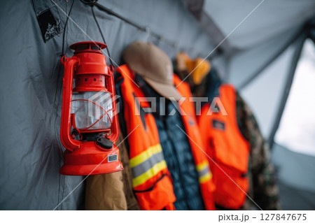 Bright red lantern hangs beside vibrant safety vests and a hat inside a camping tent, showcasing essential outdoor gear for adventurers and campers in a cozy environment 127847675