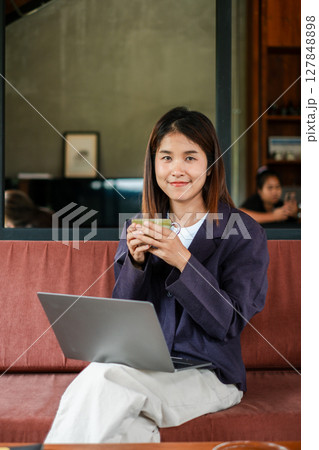 Smiling woman holding coffee cup, working on laptop in a cozy cafe. Relaxed and productive atmosphere. 127848898