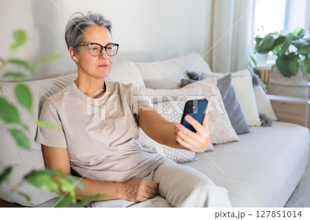 Senior woman using smartphone with glasses on, sitting on sofa in bright modern living room Senior woman using smartphone with glasses on, sitting on sofa in bright modern living room 127851014