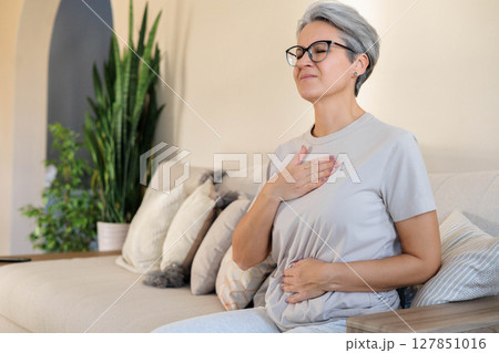 Senior woman feeling chest discomfort while sitting on sofa in cozy home environment 127851016