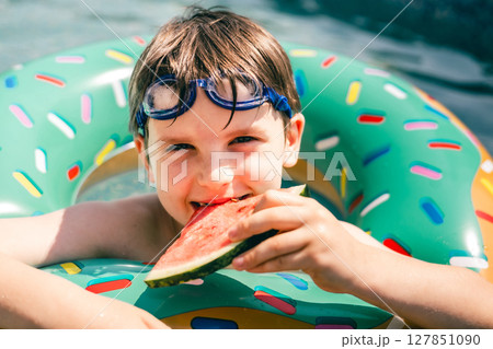 Boy biting watermelon in pool float during summer day, smiling in water with swimming goggles 127851090
