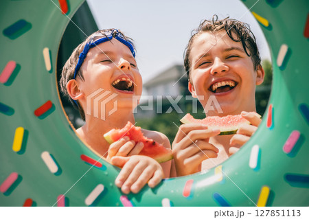 Two happy boys eating watermelon in summer through a colorful pool ring, smiling and playful Two happy boys eating watermelon in summer through a colorful pool ring, smiling and playful 127851113