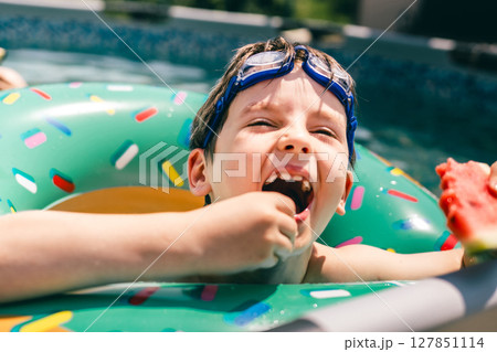 Happy boy in float eats watermelon and laughs in summer pool, enjoying sweet juicy fruit fun Happy boy in float eats watermelon and laughs in summer pool, enjoying sweet juicy fruit fun 127851114