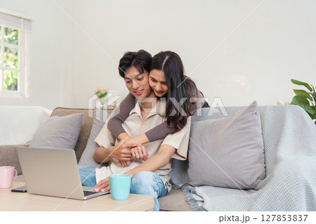 Young Couple Enjoying Quality Time Together at Home, Relaxing on the Couch with a Laptop, Smiling and Embracing in a Cozy Living Room Setting, Perfect for Lifestyle and Home Comfort Themes 127853837