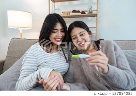 Two happy gay lesbian lgbt women couple sitting on sofa celebrating positive pregnancy test. lgbtq concept 127854083