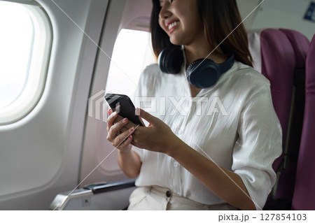 Beautiful Asian in aeroplane. working, travel, business concept Traveler on Plane Using Smartphone with Headphones, Smiling and Happy 127854103