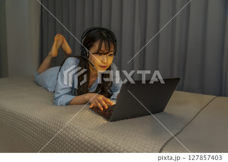 深夜にノートパソコンを操作する若い女性（ヘッドセット装着・ベッド上でリモート対応） 127857403