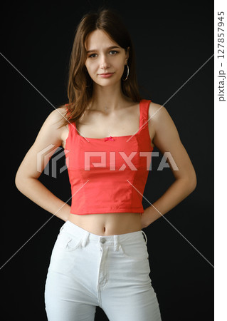 Teenager in a red top and white jeans poses confidently against a dark background 127857945