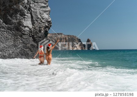 Women in Santa Claus hats run into the sea dressed in red swimsuits. Celebrating the New Year in a hot country 127858198
