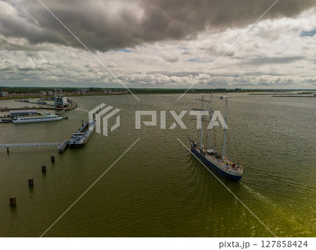A majestic sailing ship glides across calm waters under a partly cloudy sky, with coastal landscapes and a modern dam structure visible in the background. A majestic sailing ship glides across calm waters under a partly cloudy sky, with coastal landscapes and a modern dam structure visible in the background. 127858424
