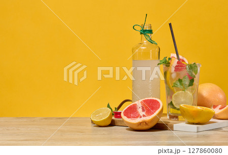 Fresh citrus drink with lemon, grapefruit, and mint in a glass with ice cubes and a straw. Surrounded by fresh citrus fruits and a bottle of homemade lemonade. 127860080