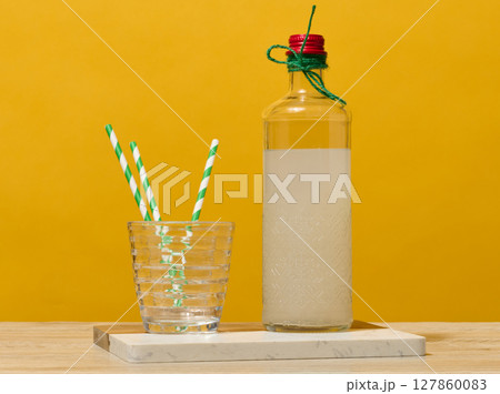 A transparent glass bottle of lemonade stands next to a faceted glass containing three green striped straws. A transparent glass bottle of lemonade stands next to a faceted glass containing three green striped straws. 127860083
