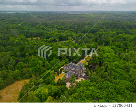 An aerial view of a historic mansion surrounded by lush green trees and a beautifully landscaped garden with a fountain, walking paths, and parking area. The scene shows vibrant summer greenery. 127860431