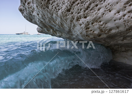 Powerful wave hitting under limestone overhang with sailboat on bright turquoise sea Powerful wave hitting under limestone overhang with sailboat on bright turquoise sea 127861617