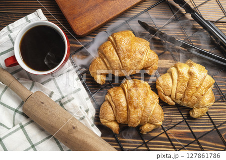 Rolling pin and tongs with freshly baked croissant on a cooling tray and a cup of coffee with a wooden background. Top view 127861786