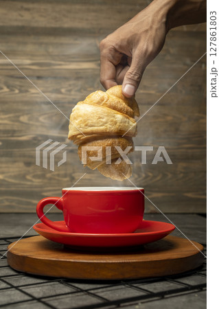 Closeup view human hand dipping a freshly baked croissant into a cup of coffee on a wooden tray with a wooden background 127861803