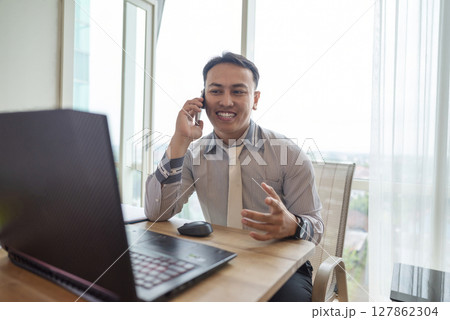 Indonesian southeast asian businessman working with a laptop while talking on the cellphone with a partner inside the hybrid workplace. Business concept. Remote working from home 127862304