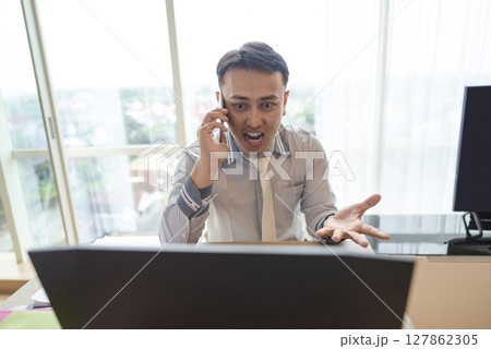 Indonesian southeast asian businessman working with a laptop while talking on the cellphone with a partner inside the hybrid workplace. Business concept. Remote working from home 127862305