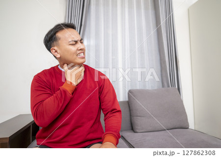 Indonesian southeast asian man sitting on the couch at home and coughing. Suffers from an attack of asthma or allergies. Healthcare concept Indonesian southeast asian man sitting on the couch at home and coughing. Suffers from an attack of asthma or allergies. Healthcare concept 127862308
